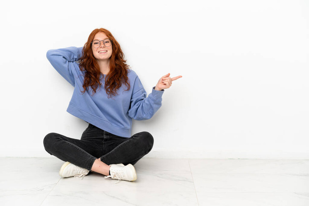 Teenager redhead girl sitting on the floor isolated on white background surprised and pointing finger to the side - Foto, Imagen