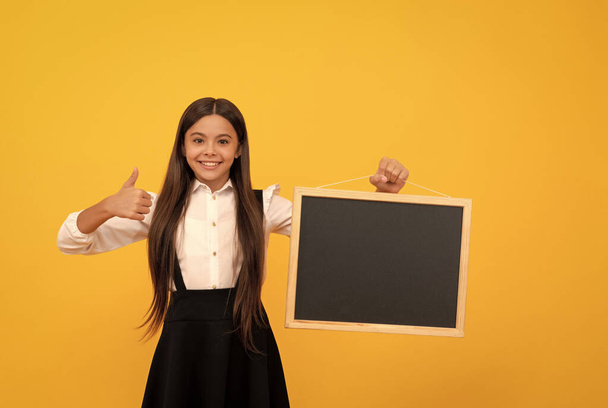 wesoły nastolatek dziewczyna w mundurze trzymać szkoła tablicy dla ksero przestrzeń pokazując kciuk w górę, prezentując - Zdjęcie, obraz