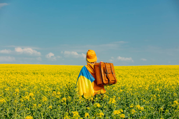 Γυναίκα με κίτρινη κουκούλα με ουκρανική σημαία και βαλίτσα στο χωράφι με τους κραμβόσπορους - Φωτογραφία, εικόνα