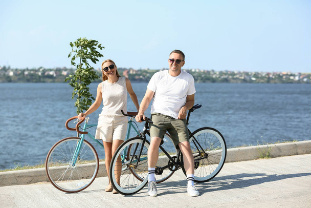 Mature couple with bicycles walking along river bank on summer day - Photo, Image