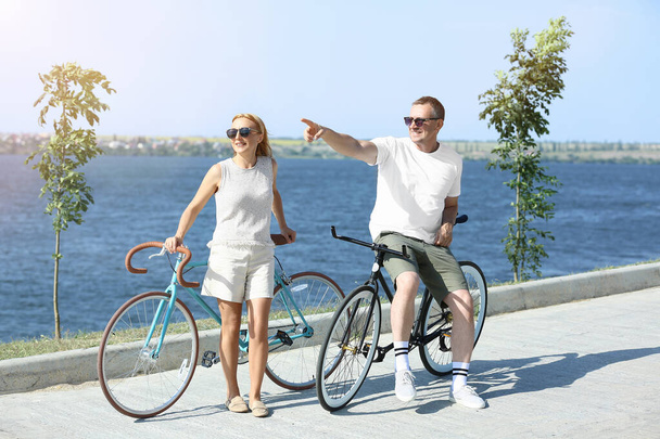 Pareja madura con bicicletas caminando a lo largo de la orilla del río en el día de verano - Foto, Imagen