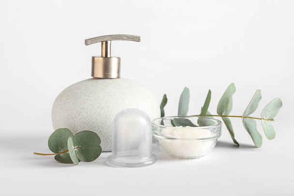 Bottiglia di prodotto cosmetico, ciotola con sale marino, vasetto sottovuoto per massaggio anticellulite e ramo di eucalipto su fondo bianco - Foto, immagini