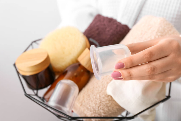 Woman holding basket with bath supplies and vacuum jars for anti-cellulite massage on light background, closeup - Photo, Image
