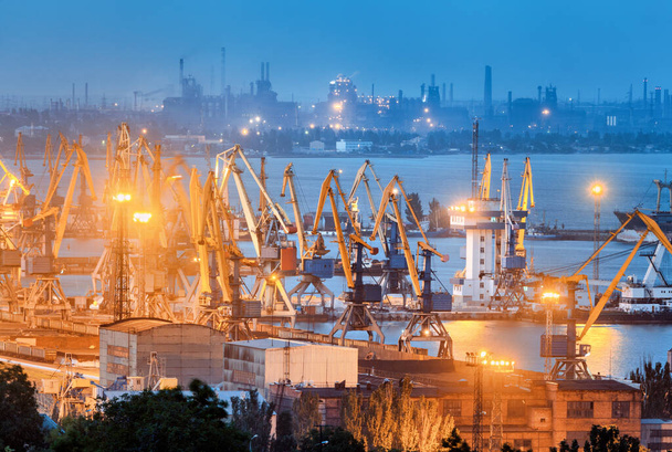 Sea commercial port and steel plant at night in Mariupol, Ukraine before the war. Industrial landscape. Cargo freight ship with working cranes bridge in sea port at dusk. Cargo port, logistic. - Foto, Imagen