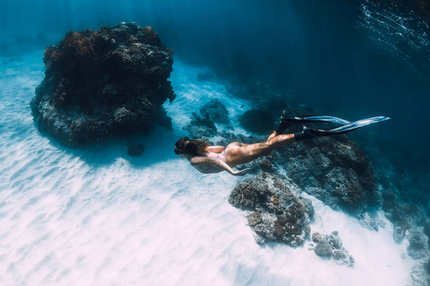 Žena v bikinách se potápí na písčitém dně s tropickými korály. Atraktivní štíhlá dívka s ploutví pod vodou - Fotografie, Obrázek
