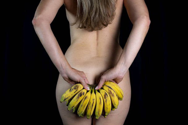 Woman with back to camera holding banana tree against black background in studio. Salvador, Bahia, Brazil. - 写真・画像