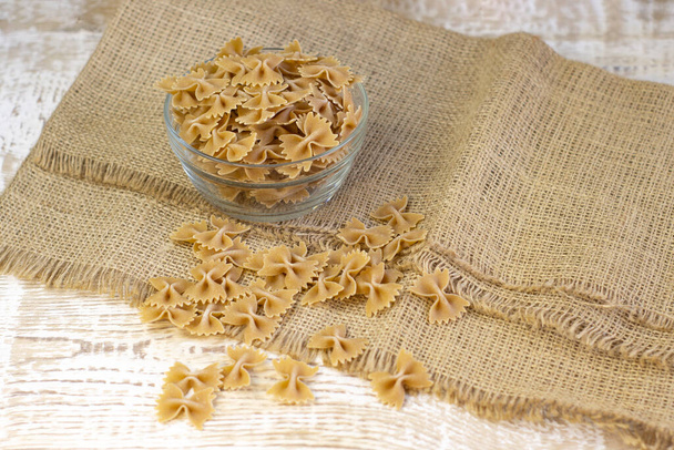 Whole grain dark pasta bows on brown plate on a rough wooden light background with selected focus - Photo, image