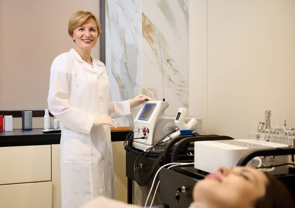Pleasant mature Caucasian woman, female beautician standing near modern medical equipment for body beauty treatment in wellness spa clinic - Foto, imagen