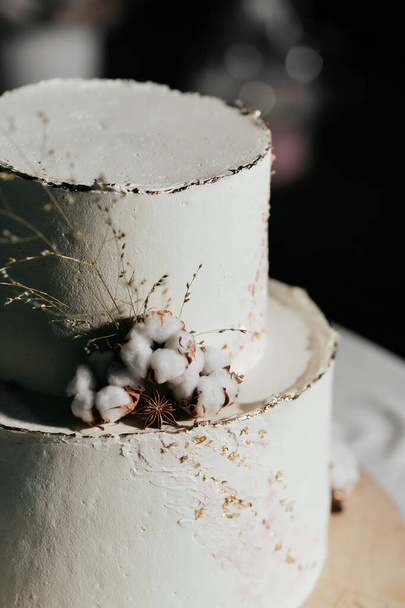 Tort weselny. Ciasto w kolorze mleka ozdobione bawełnianymi koralikami i złotym liściem na ciemnym tle - Zdjęcie, obraz