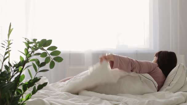 jó reggelt koncepció - fiatal nő rózsaszín pizsamában ébredés fényes hálószobában - Felvétel, videó