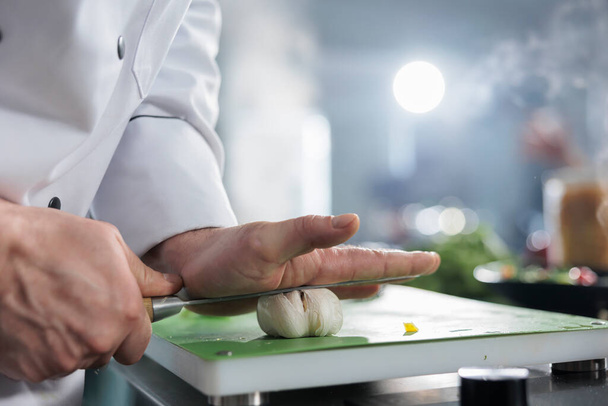 Gros plan des mains de cuisinier en chef à l'aide d'un couteau de cuisson pour casser une tête d'ail frais pour le plat de service du dîner. - Photo, image