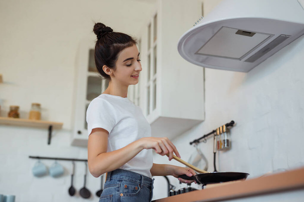 Feliz joven y atractiva cocinera ama de casa preparando la comida en una sartén en la estufa para una deliciosa cena en la cocina moderna estilo loft - Foto, Imagen