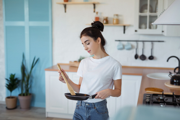 Ευτυχισμένος ελκυστική νεαρή νοικοκυρά μαγείρεμα γυναίκα προετοιμασία των τροφίμων σε ένα τηγάνι για νόστιμο δείπνο στη σύγχρονη κουζίνα στυλ σοφίτα - Φωτογραφία, εικόνα