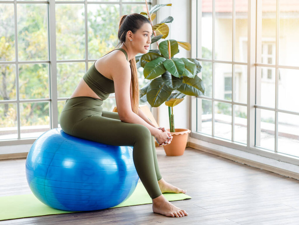 Closeup shot of Millennial Asian young healthy slim strong sexy female sporty model in casual crop top sportswear sitting on exercise ball smiling posing holding hand at chin in classroom gym studio. - 写真・画像