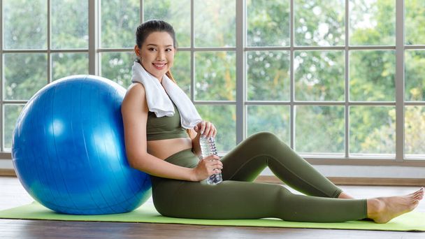 Closeup shot Millennial Asian young healthy slim strong sexy female sporty model in casual crop top sportswear sitting resting relaxing leaning on exercise ball drinking water in bottle in classroom. - Фото, изображение