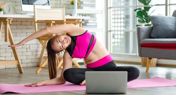 Asian young beautiful sexy slim strong female sporty model in crop top sportswear sitting smiling stretching crossed legs and arms cool down on yoga pilates mat in apartment living room after workout. - Fotó, kép