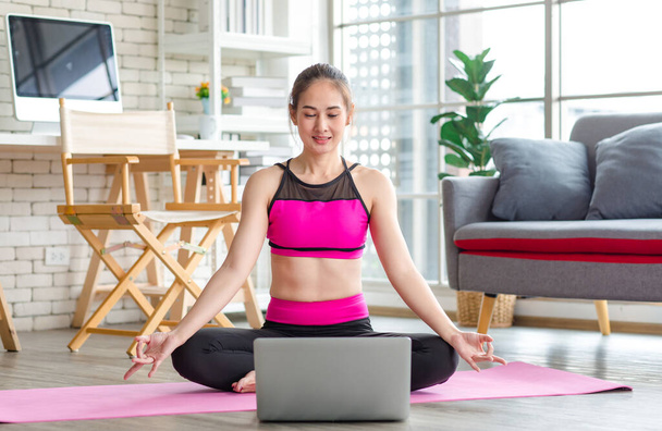 Asian young beautiful sexy slim strong female sporty model in crop top sportswear sitting smiling stretching crossed legs and arms cool down on yoga pilates mat in apartment living room after workout. - Фото, изображение