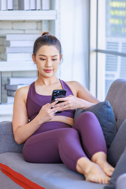 Portrait shot Millennial Asian young healthy slim strong sexy female sporty model in casual crop top sportswear laying down smiling look at camera on cozy sofa in living room using smartphone online. - 写真・画像
