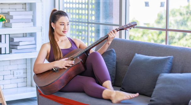 Millennial Asian young beautiful healthy slim strong sexy female sporty model in casual crop top sportswear sitting smiling holding playing acoustic guitar on cozy sofa couch in living room at home. - Photo, Image