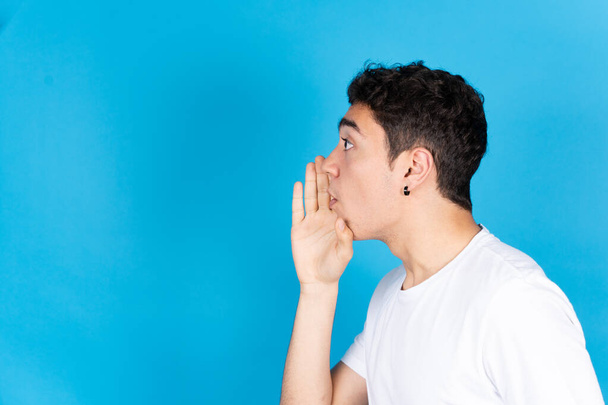 Side view of hispanic teenager boy telling a secret hand on mouth to copy space isolated on blue background. - Foto, Imagen