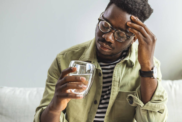Young man suffering from strong headache or migraine sitting with glass of water in the living room, millennial guy feeling intoxication and pain touching aching head, morning after hangover concept - Foto, imagen