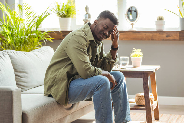 Frustrated middle aged man with hand on head sitting on couch at home. Worried African American business man with terrible migraine, overload and overworked concept. - Photo, Image