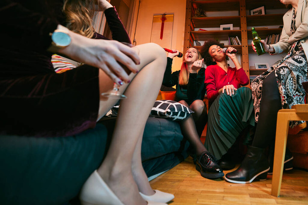Wielokulturowa grupa przyjaciółek śpiewa karaoke podczas picia wina w salonie. - Zdjęcie, obraz