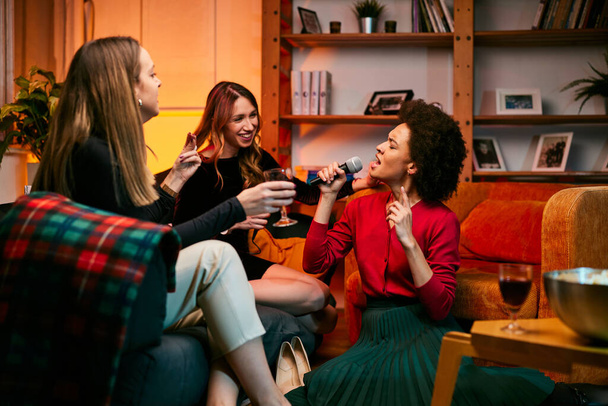 Çok kültürlü bir kadın arkadaş grubu oturma odasında şarap içerken karaoke yapıyor.. - Fotoğraf, Görsel