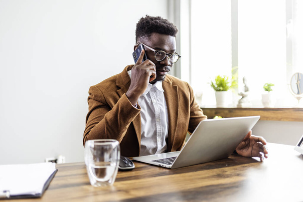 confident african american businessman sitting at desk with laptop talking on phone  - Fotó, kép