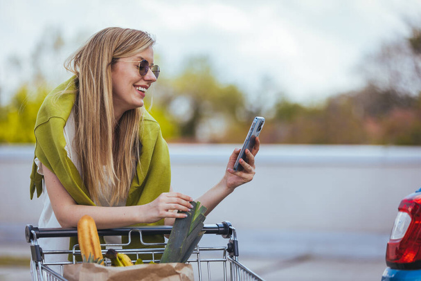 young woman with shopping cart and smartphone standing near car - Foto, Bild