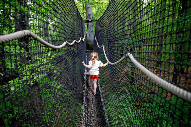 Linda niña preescolar caminando por el sendero alto del dosel del árbol con pasarela de madera y cuerdas en Hoherodskopf en Alemania. Feliz niño activo explorando el camino de la copa de un árbol. Actividad divertida para familias al aire libre - Foto, imagen