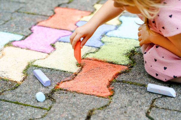 裏庭の地面にカラフルなチョークで小さな就学前の女の子の絵。肯定的な幸せな幼児の子供の絵を描き、絵を作成する。夏の創造的な屋外活動. - 写真・画像