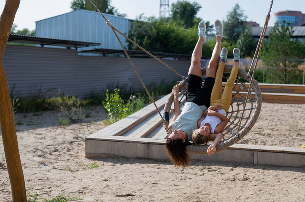 Mom and daughter swing on a round swing. Caucasian woman and little girl have fun on the playground. - Foto, Imagem