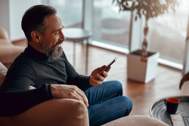Smiling bearded man sitting in armchair and reading message on smartphone - Foto, Bild
