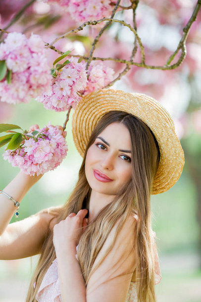 Spring portrait of a young beautiful woman in pink blossoms. Young beautiful model with long hair, in a wicker hat and a light dress near the cherry blossoms. Spring. - Photo, Image