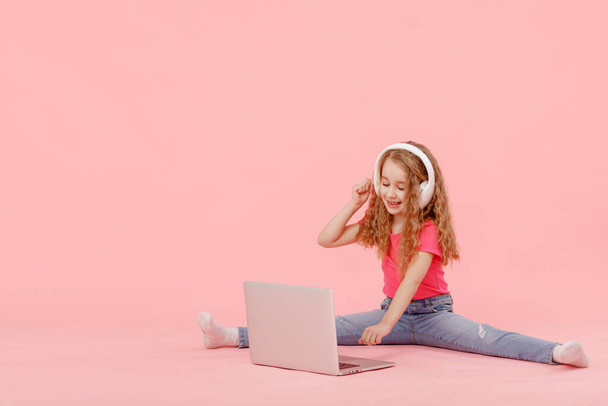adorable child girl is sitting in the twine next to a laptop. kid in white headphones stretching and learns homework or watching cartoons, listening music. - Zdjęcie, obraz