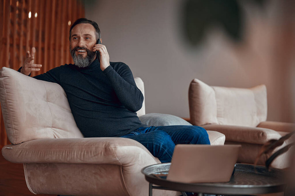Smiling bearded man sitting in armchair and chatting on phone - Valokuva, kuva