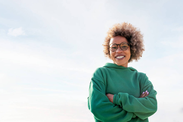 young latin woman with glasses and afro hair smiling happy looking at camera with arms crossed and the sky in the background, concept of happiness and lifestyle, copy space for text - Foto, immagini