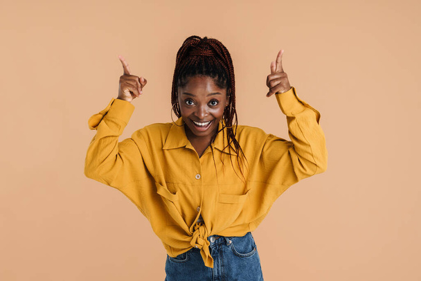 Young black woman with afro pigtails smiling and pointing fingers upward isolated over beige background - Fotó, kép