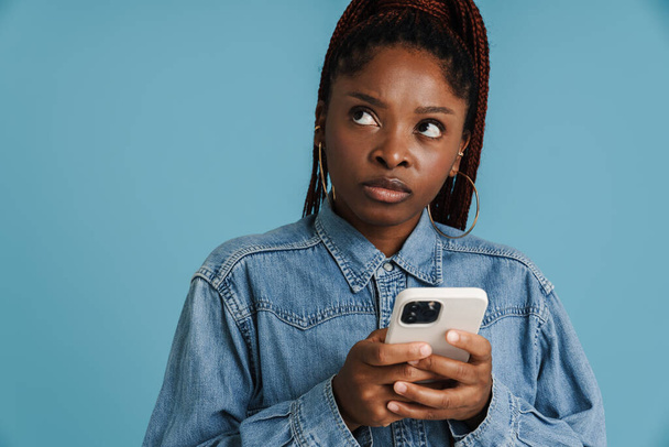 Νεαρή μαύρη γυναίκα με αφρικανικές κοτσίδες χρησιμοποιώντας κινητό τηλέφωνο απομονωμένο σε μπλε φόντο - Φωτογραφία, εικόνα