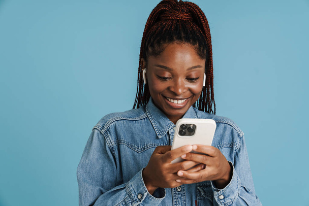Young black woman smiling while using cellphone and earphones isolated over blue background - Foto, Imagem