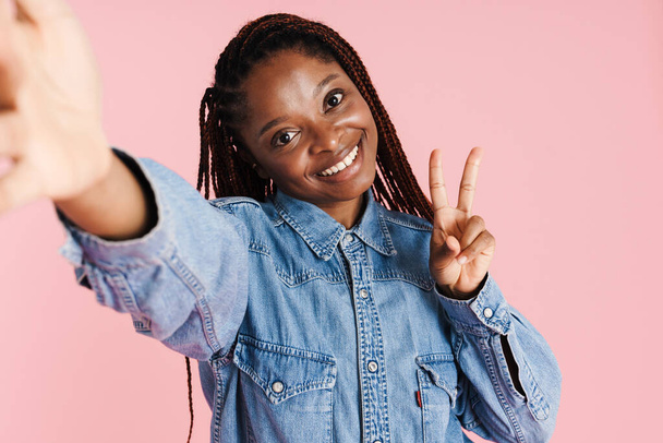 Young black woman showing peace sign while taking selfie photo isolated over pink background - Foto, afbeelding