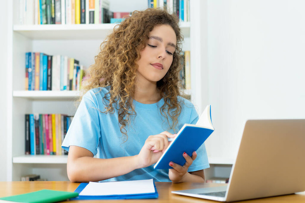 Pretty caucasian female student reading reference book at desk at university - Zdjęcie, obraz