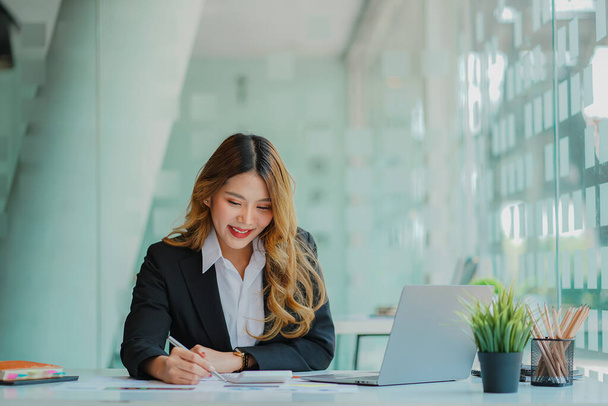 An Asian female accountant sits at a desk with a laptop and calculates financial graphs showing investment results. Plan a successful business growth process in the office. - Zdjęcie, obraz
