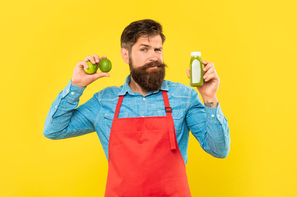 Winking man in apron holding limes and juice bottle yellow background, juice barman - Foto, Bild