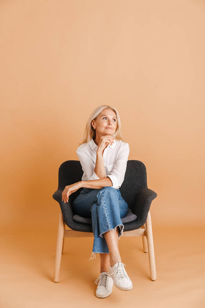 Mature blonde woman looking aside while sitting in chair isolated over beige background - Foto, Bild