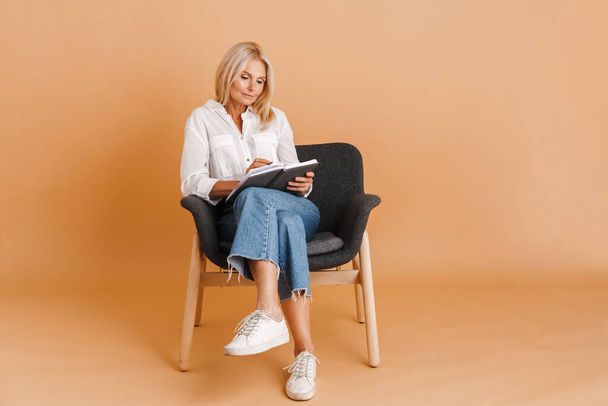 Mature blonde woman writing down notes while sitting in chair isolated over beige background - Foto, Imagem