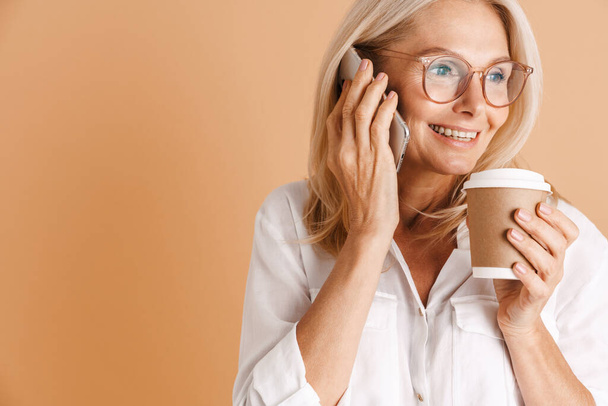 Mature woman talking on cellphone while drinking coffee isolated over beige background - Фото, зображення