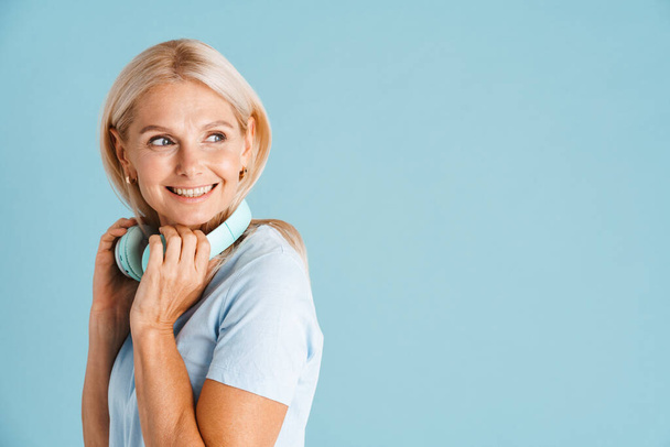 Mature blonde woman with headphones smiling and looking aside isolated over blue background - Zdjęcie, obraz