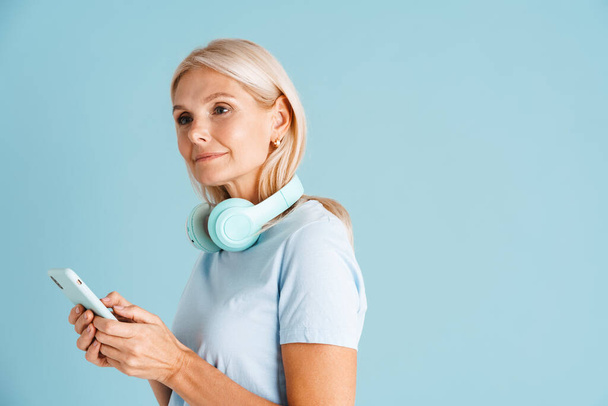 Mature blonde woman with headphones using mobile phone isolated over blue background - Φωτογραφία, εικόνα